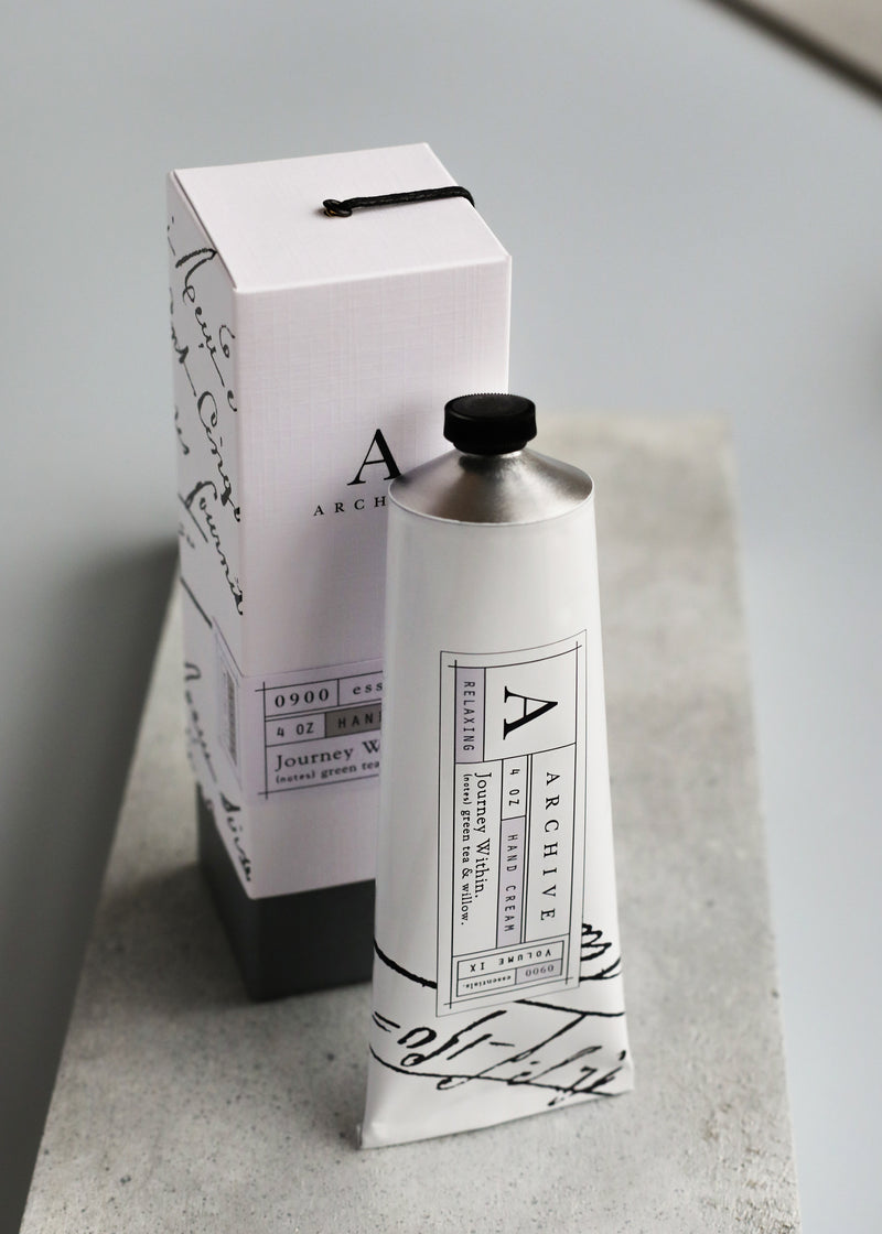 A metal tube of ARCHIVE by Margot Elena - Journey Within Hand Cream next to its packaging box on a gray surface. The tube and box feature a minimalistic black and white design with elegant typography.