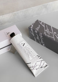 A stylishly designed metal tube labeled "ARCHIVE by Margot Elena - Journey Within Hand Cream" on a gray surface next to a book with a pink cover and another wrapped in a black-and-white patterned paper