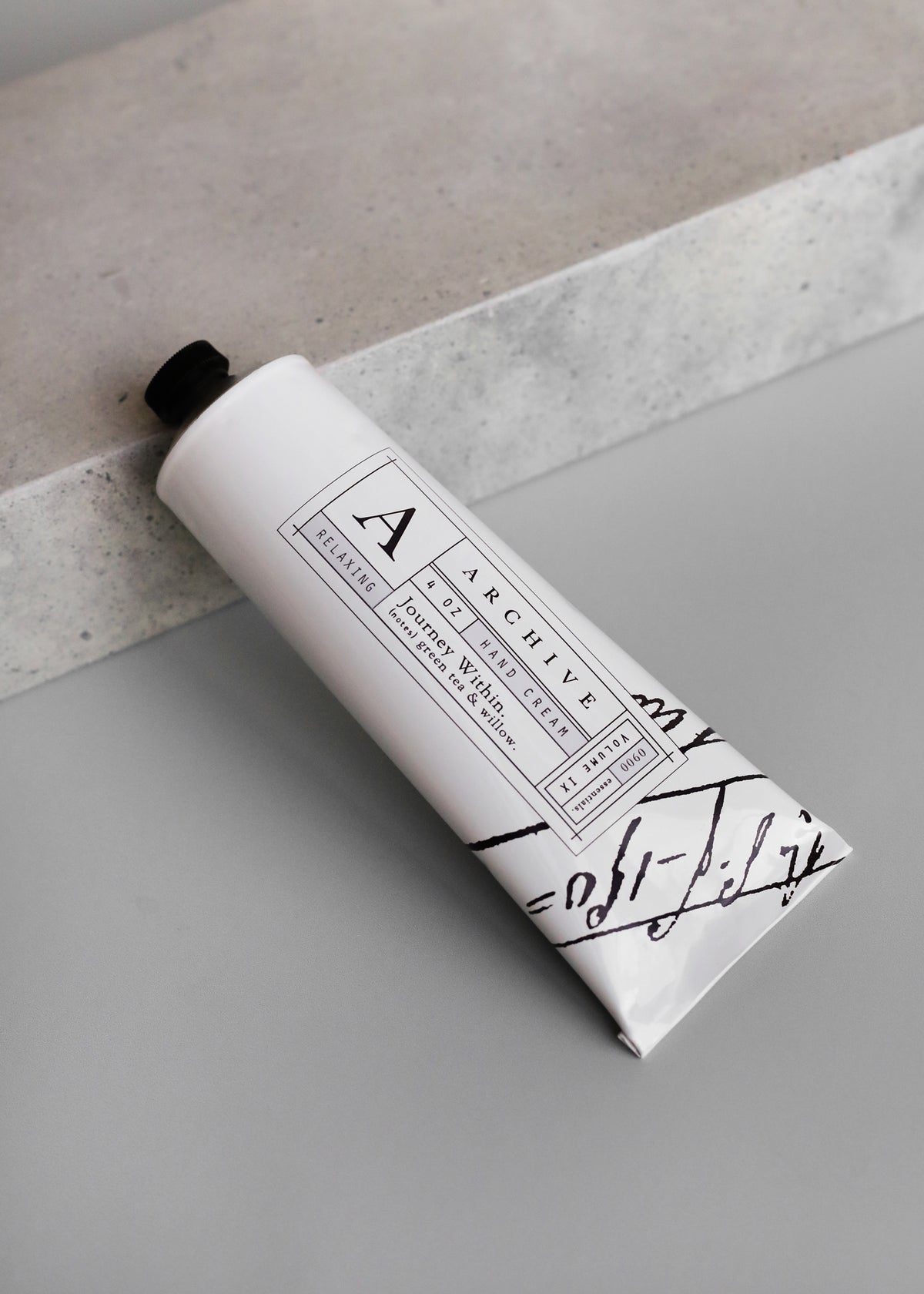 A tube of ARCHIVE by Margot Elena - Journey Within hand cream with a minimalistic black and white design resting on a gray stone surface.