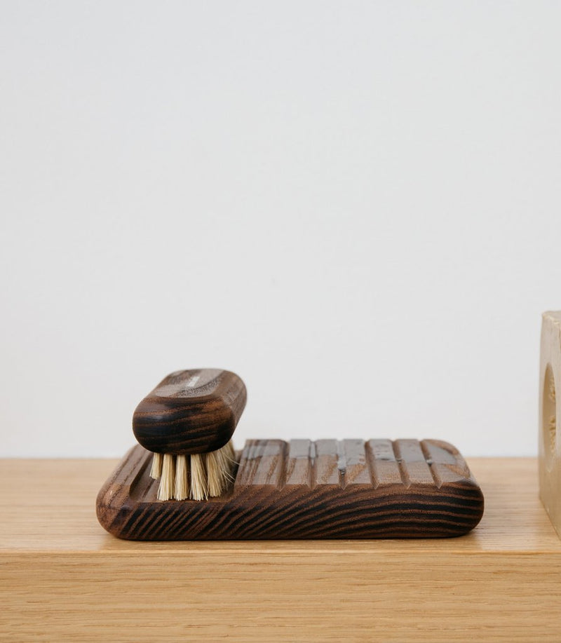 A dark wooden scrubbing brush resting on an Andrée Jardin Heritage Ash Soap Holder with firm bristles, set against a neutral background, sitting on a light wooden surface.