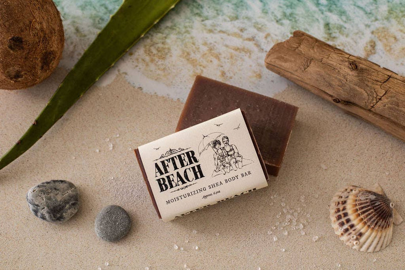 A bar of Primitive House Farm - After Beach Shea Butter Soap placed on a sandy surface surrounded by a coconut, aloe vera leaf, driftwood, pebbles, and a seashore.