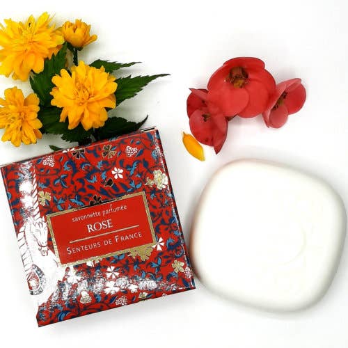 A flat lay image of a Senteurs De France Rose-scented soap "Unicorn" with organic Shea Butter in a decorative red and blue box, alongside fresh yellow and red flowers and a white ceramic dish.