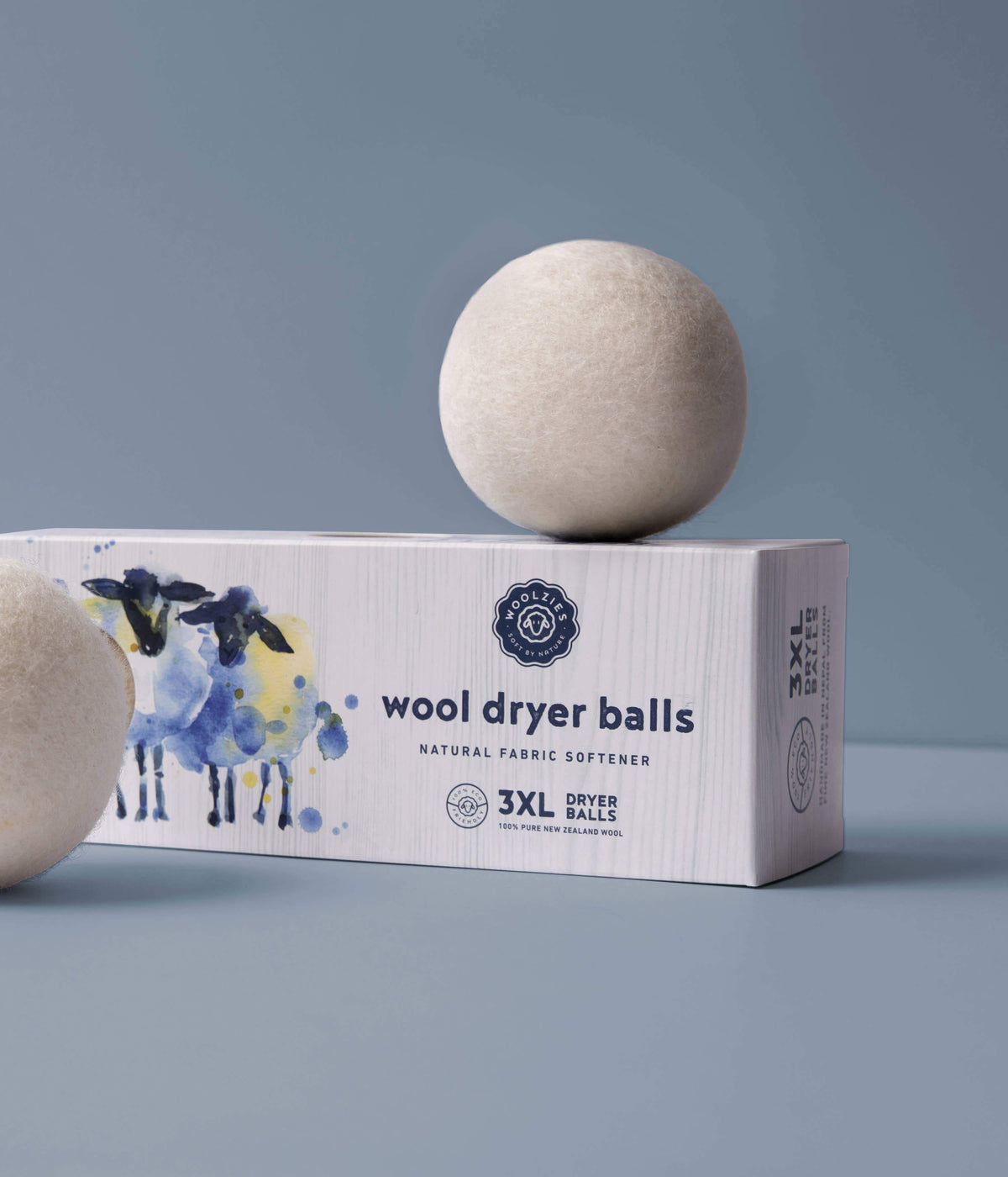 A box labeled "Woolzies Wool Dryer Balls - Set of 3" with a watercolor image of sheep on the side, with two wool balls resting beside and on top of it against a gray background.