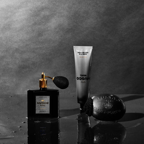 Black-and-white image of luxury grooming products including a perfume bottle with a spray bulb, a tube of Claus Porto 1887 Musgo Real Black Edition Shaving Cream, and a round soap, all arranged on a reflective surface with water droplets.