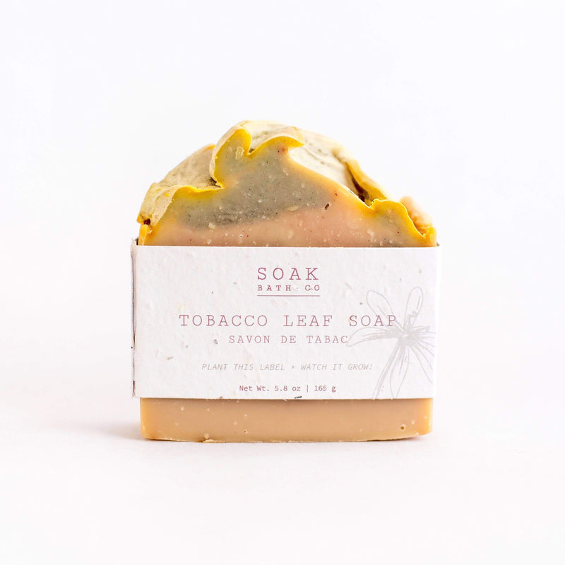 A bar of SOAK Bath Co. - Tobacco Leaf Soap, featuring a layered design with a yellow top and beige bottom, wrapped in a white label with plant illustrations.