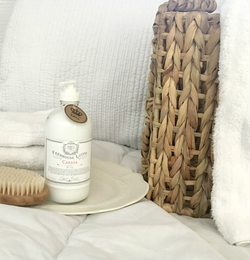 A cozy home setting featuring a bottle of Z&Co. Cabana Farmhouse Lotion, enriched with Shea Butter, a white plate, and a brush beside a woven pillow on a white textured couch.