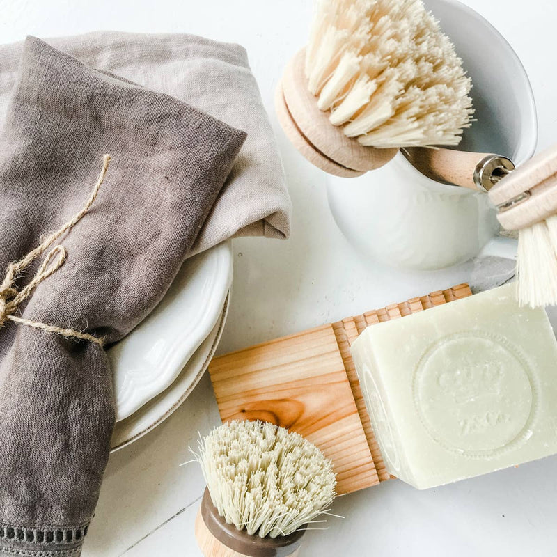 A variety of eco-friendly cleaning tools: brushes with wooden handles, a bar of soap on a Z&Co. Cedar Wood Block Soap Holder, stack of plates, and a linen napkin, all arranged on a white surface.