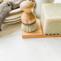 A Z&Co. cedar wood block soap holder with a round handle next to a large block of pale green farmhouse kitchen soap, positioned beside stacked ceramic plates on a white surface.
