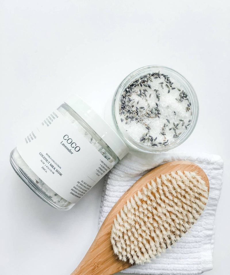 A top view of two jars of Z&Co. brand paraben-free skincare products next to a wooden dry brush on a white towel, with one jar open displaying Z&Co. Coco + Lavender Coconut Milk Soak sprinkled with herbs.