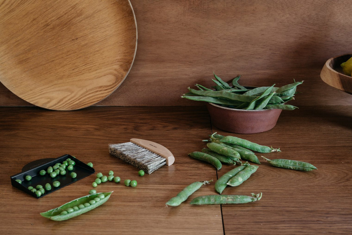 A wooden table with fresh peas, some still in their pods, next to a bowl of pea pods, an Andrée Jardin Mr. & Mrs. Clynk Natural Table Brush & Dustpan Set - Black, and a wooden plate.
