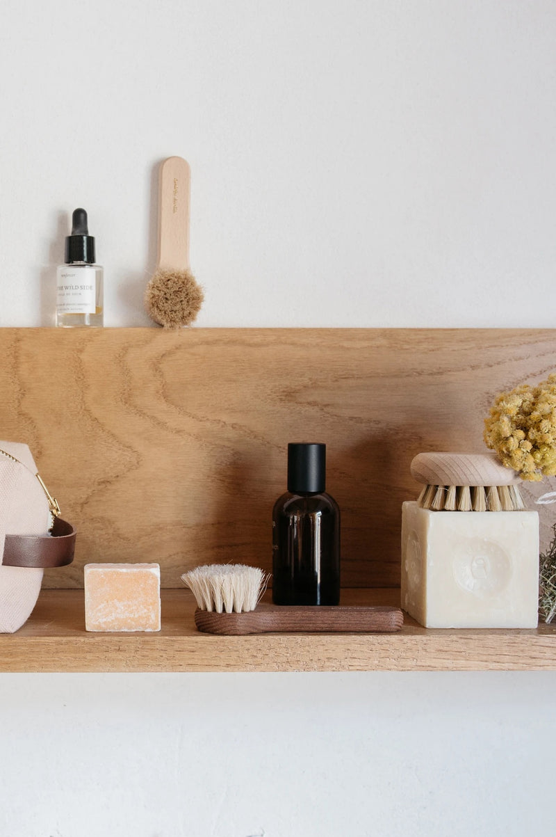 A minimalist wooden shelf displaying assorted self-care items including an Andrée Jardin Tradition Face Cleansing Brush Waxed Beech Wood, bottles, soap bars, and a dried flower bundle against a white wall.