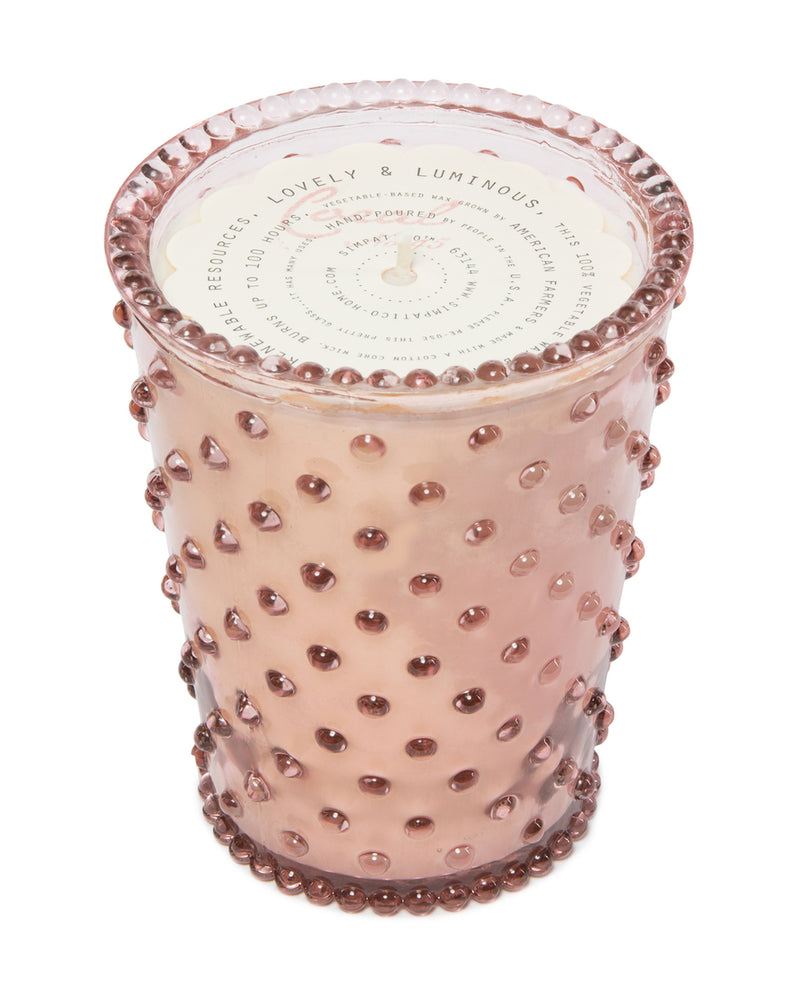 A pink Simpatico NO. 45 CORAL Hobnail Glass Candle holder with a textured surface featuring raised dots, and a rim designed with a scalloped pattern. The lid's label reads "sunny citrus & luminous.