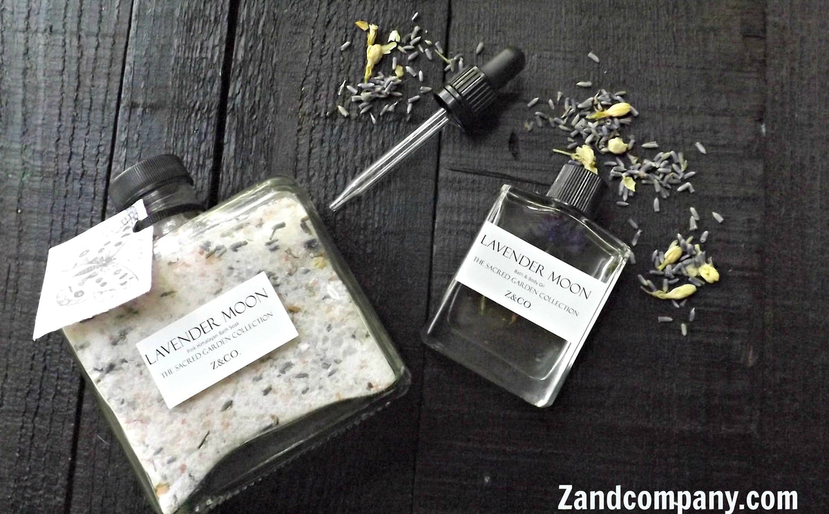 Two Z&Co. Pink Himalayan Bath Soak Lavender Moon glass bottles containing bath soak and essential oil, with dried lavender scattered around, on a dark wooden surface.