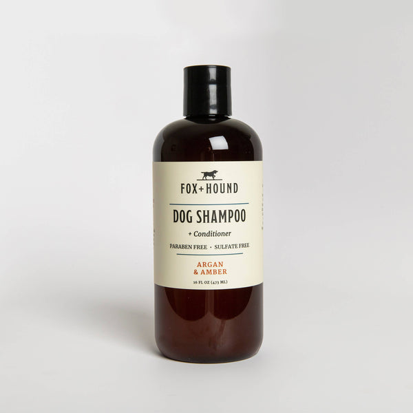 A brown bottle of "Fox + Hound Argan & Amber Dog Shampoo + Conditioner" against a white background. The label mentions 'paraben free, sulfate free' with key ingredients like 'argan