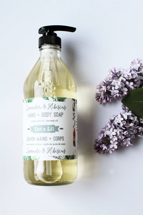 A clear bottle of Dot & Lil Lavender & Hibiscus hand and body soap, placed on a white surface next to fresh purple lilacs. The bottle features a pump top and descriptive labels highlighting its pure so