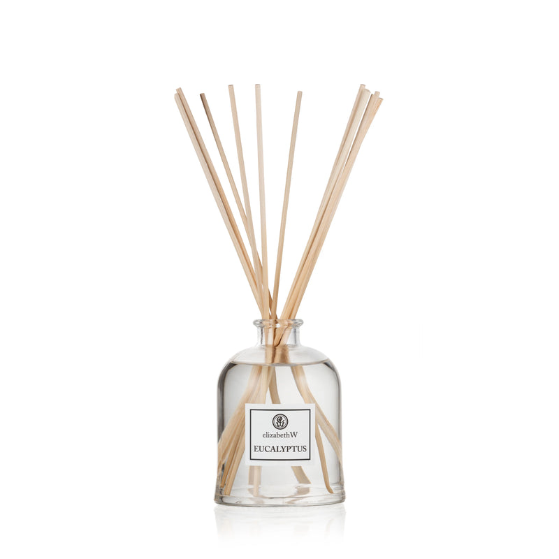 A clear glass environmentally friendly diffuser bottle labeled "Elizabeth W Purely Essential Eucalyptus Diffuser" with several light brown reed sticks inserted, isolated on a white background.