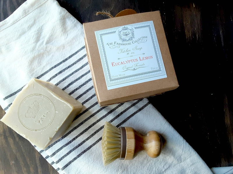 A Z&Co. Farmhouse Solid-Block Eucalyptus Lemon Dish Soap next to its open packaging box and a wooden brush on a striped cloth on a dark wooden surface.