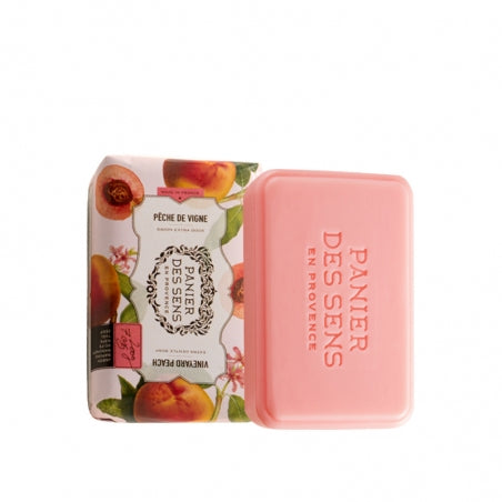 A Panier des Sens Extra-Soft Vegetable Soap - Vineyard Peach, infused with shea butter, next to its packaging, which has images of peaches and French text on it, against a white background.