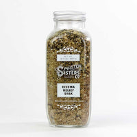 A clear glass jar filled with a mixed herbal blend labeled "Spinster Sisters Co. Eczema Relief Soak." The jar contains colloidal oatmeal and is sealed, displaying product details on a card attached to the lid.