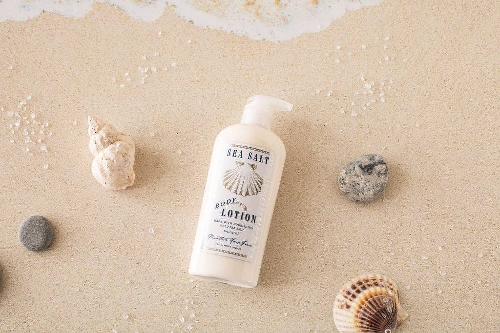 A bottle of Primitive House Farm - Sea Salt Lotion lies on a sandy surface, surrounded by various seashells, evoking a beach-like atmosphere.