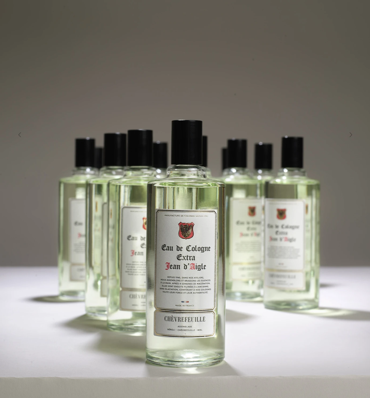A collection of glass bottles filled with Jean d'Aigle Honeysuckle Eau De Cologne, labeled "Jean d'Aigle" with some bottles slightly out of focus in the background, centered on a well.