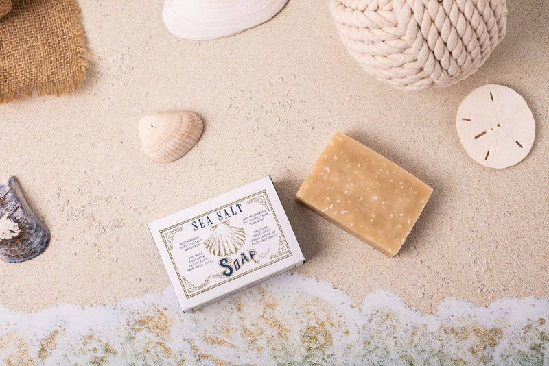 A bar of Primitive House Farm - Sea Salt Soap next to its packaging, surrounded by seashells, a rope, and a wave of foam on a sandy background, evoking a beach setting.