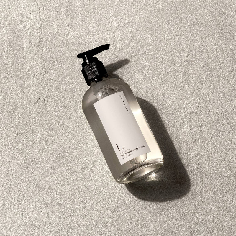A clear glass bottle of Lavande Lavender Body Wash sits on a textured gray surface, casting a soft shadow. The label is minimalistic, featuring simple black text on the organic base soap.
