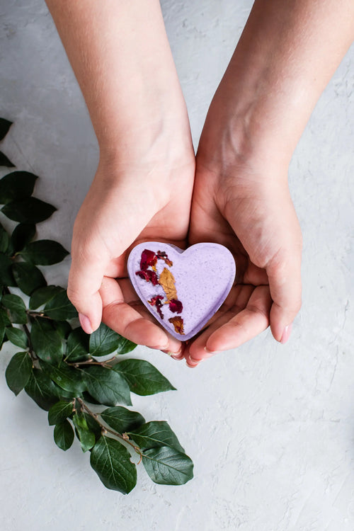 Two hands cradling a Lizush Lavender Heart Shower Steamer bath bomb with flower petals and essential oils, positioned above a white surface with green leaves around.
