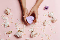 Hands holding a purple, heart-shaped sponge surrounded by scattered white and pink rose petals and a Lizush Lavender Heart Shower Steamer on a soft pink background.