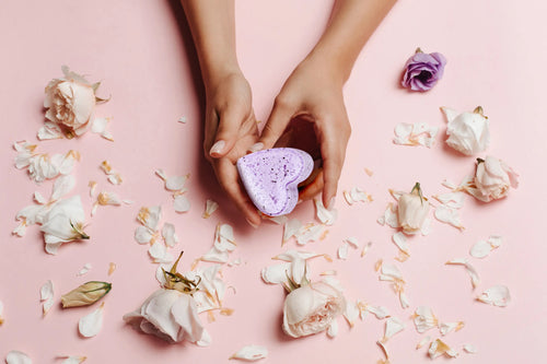 Hands holding a purple, heart-shaped sponge surrounded by scattered white and pink rose petals and a Lizush Lavender Heart Shower Steamer on a soft pink background.