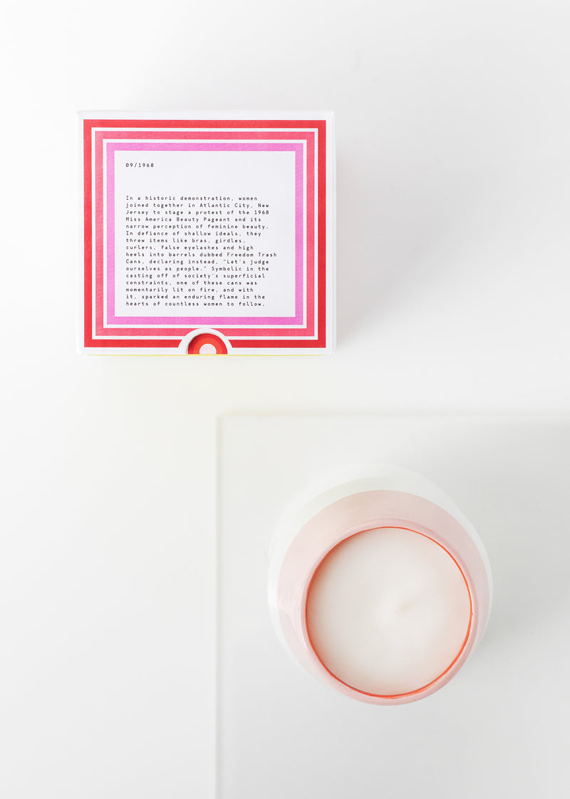 A top view of a white surface with a pink and white book next to a Margot Elena Infinite She Burn Baby Burn Ceramic Candle in a white and pink bowl. The book is open to a page with text.