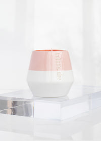 A Margot Elena Infinite She Burn Baby Burn Ceramic Candle with a pink and white gradient design and a reflective logo, displayed on a translucent acrylic surface against a softly lit background, infused with the scent of Fresh Cut Iris.