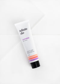 A tube of Margot Elena's "Infinite She Inspired Ultra-Lush Hand Cream" lying on a white background with a diagonal shadow line across the surface. The tube features minimalist design and pastel stripes.