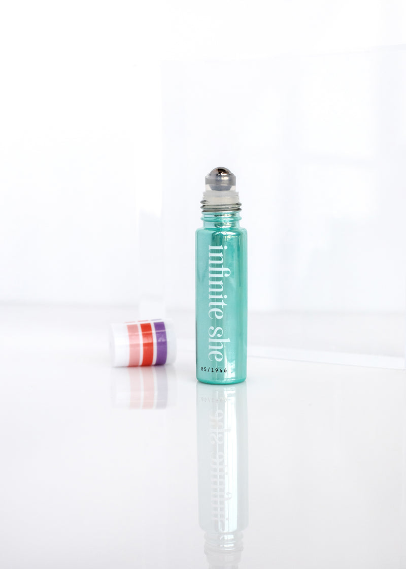 A Margot Elena Infinite She Inspired Rollerball Eau de Parfum with turquoise liquid and a silver cap, standing upright on a reflective white surface, with a blurred lip balm in the background.