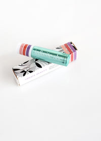 A tube of Margot Elena's Infinite She Inspired Rollerball Eau de Parfum in a teal container with colorful stripes, lying on a stylish black and white patterned box with the text "create beauty." The background is a plain white surface.