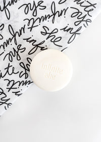 A Margot Elena soap product with "Infinite She One Badass Babe Shea Butter Soap" printed on the lid, resting on a corner of paper featuring artistic black script handwriting on a white background.