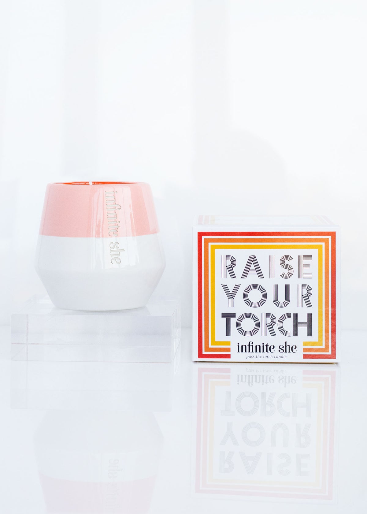 A white and peach-colored sandalwood-scented candle labeled "Infinite She Raise Your Torch Ceramic Candle" next to a cardboard box with the words "raise your torch" on a reflective white surface.