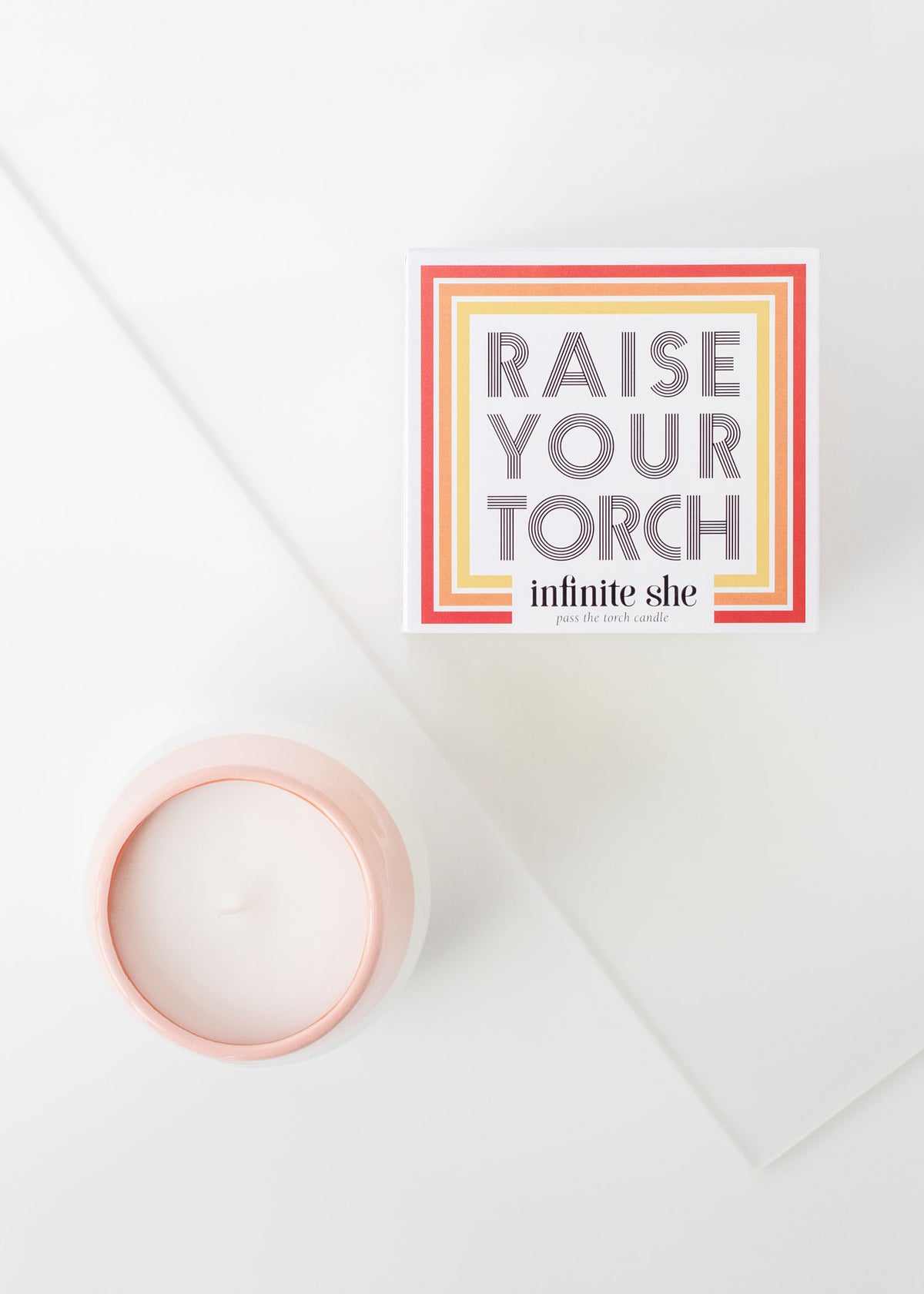A book titled "Raise Your Torch" by Infinite She lies next to a Margot Elena Infinite She Raise Your Torch Ceramic Candle infused with sandalwood on a white surface, illuminated by natural light from the left side.