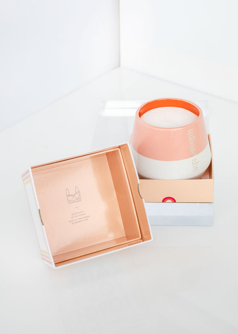 A peach-colored Margot Elena Infinite She Raise Your Torch Ceramic Candle container, infused with sandalwood, placed beside its open packaging, reflecting a simple, elegant design against a white background.