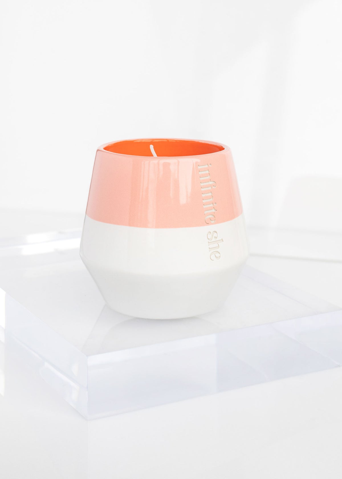 A modern, two-tone Infinite She Raise Your Torch Ceramic Candle in a white and coral container labeled "Margot Elena" on a transparent surface with a white backdrop, crafted from a Soy + Coconut Wax Blend.