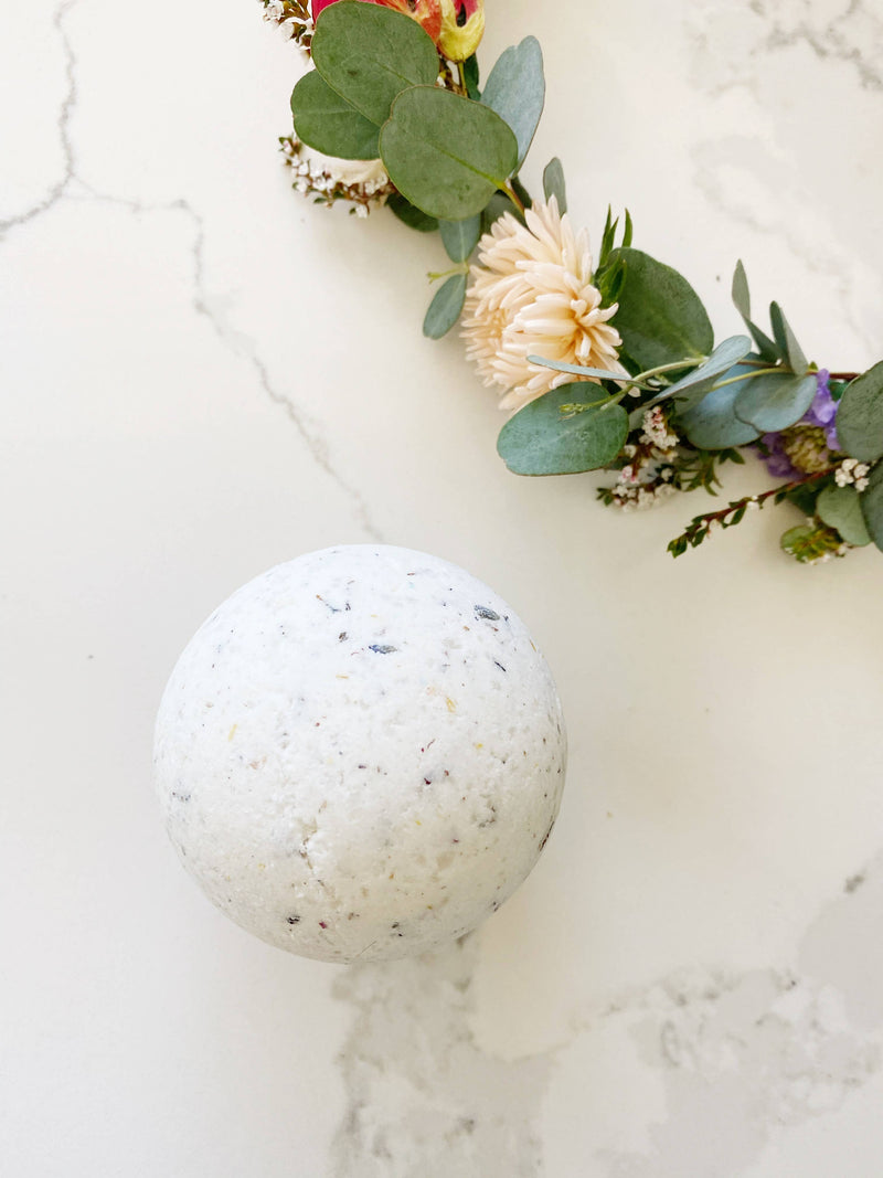 A white speckled SOAK Bath Co. - Jasmine bath bomb is placed on a marble surface, surrounded by a partial wreath of delicate flowers and greenery. The setting conveys a sense of relaxation and spa-like tranquility.