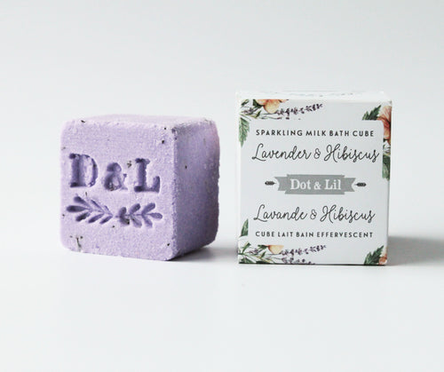 A purple sparkling bath cube with "d&l" engraved next to its packaging labeled "Dot & Lil Lavender & Hibiscus Sparkling Milk Bath Cube." The background is plain white.