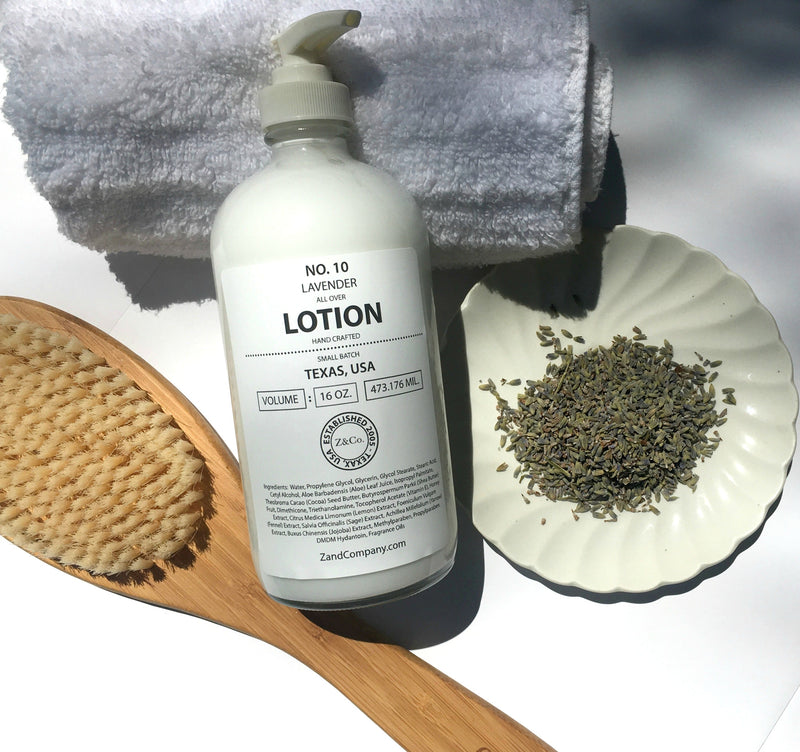 A bottle of Z&Co. Lavender Lotion 16 oz. labeled "no. 10 Lavender Lotion, Texas, USA" beside a brush, a shell-shaped dish of lavender buds, and a white towel on a