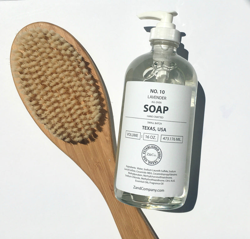A dry brush and a glass bottle of Z&Co. All Over Soap 16 oz. Lavender Body Wash And Liquid Soap labeled "no. 10 lavender soap, 16 oz, texas, usa" on a white background.