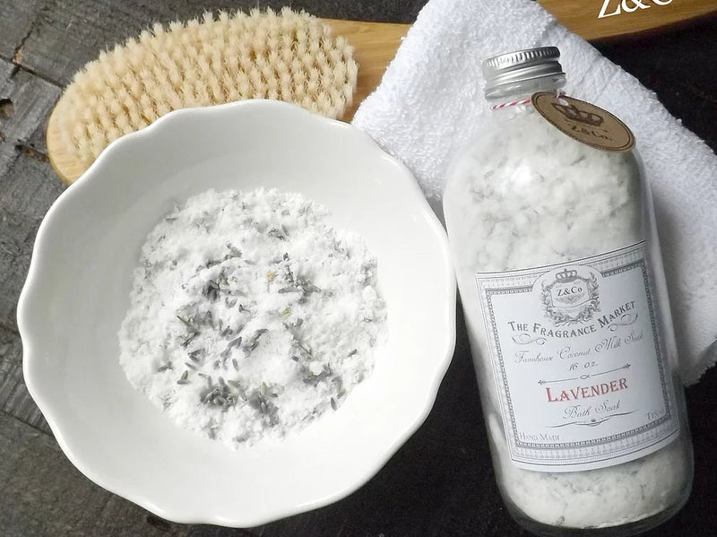 A spa setup featuring a bowl of Z&Co. Lavender 16 oz. Coconut Milk Soak bath salts with lavender buds, a dry brush, a white towel, and a bottle of lavender bath oil on a wooden surface.