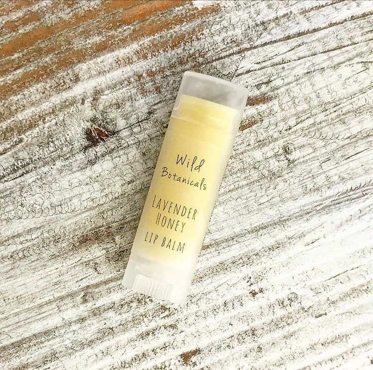 A tube of "Wild Botanicals Lavender Honey Lip Balm" by Wild Botanicals lies on a weathered wooden surface. The balm texture is visible at the top of the tube.