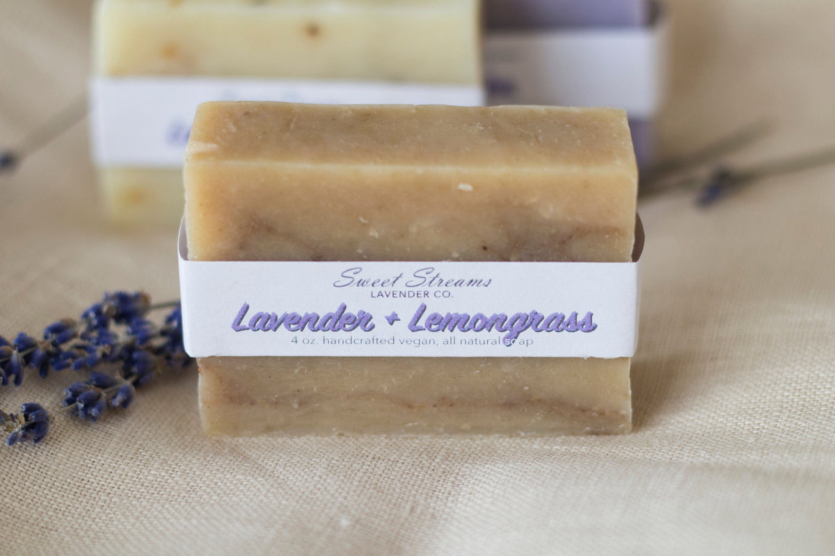 Handcrafted natural vegan soap bars labeled "lavender + lemongrass" by Sweet Streams Lavender Co., displayed with dried lavender sprigs on a textured beige surface.