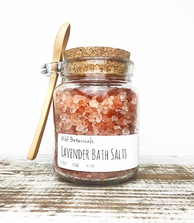 A Wild Botanicals Lavender Himalayan Bath Salts Cork Jar with pink crystals visible through the glass, complete with a cork top and a wooden spoon attached to the side. The label reads "Wild Botanicals Lavender".
