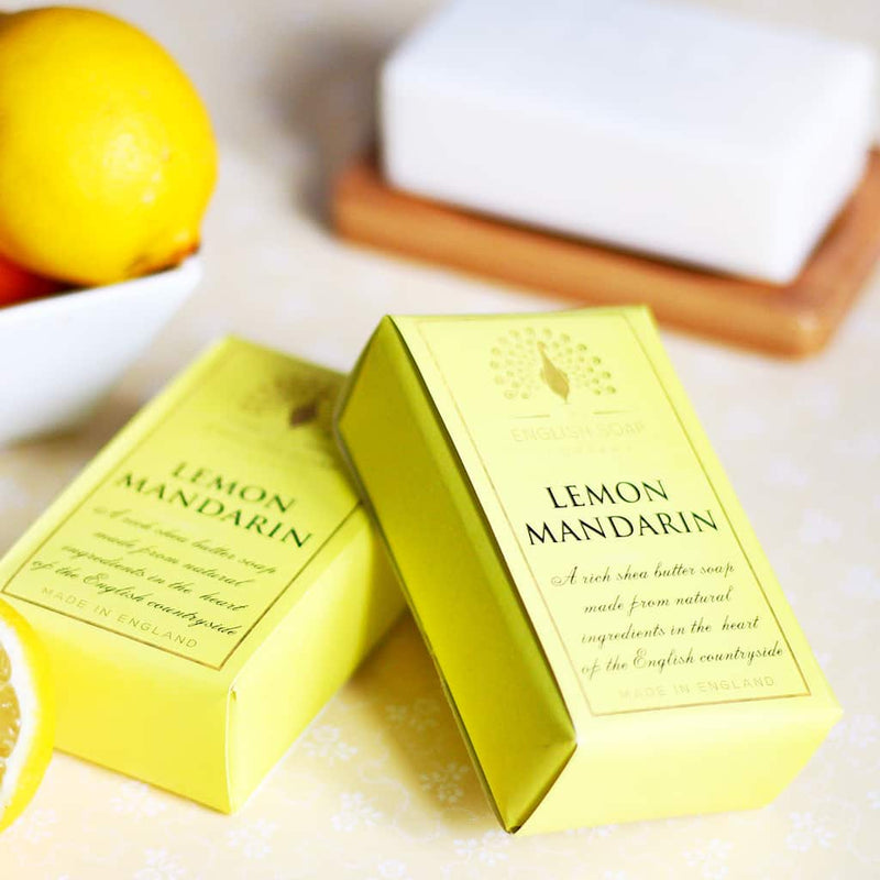 Two boxes of The English Soap Co. Pure Indulgence Lemon Mandarin soap on a kitchen counter, with a lemon, a white dish, and a subtle floral background. One box is open, displaying a yellow soap bar.