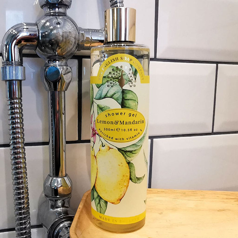 A The English Soap Co. Vintage Lemon & Mandarin Shower Gel bottle, enriched with vitamin E, stands beside a silver sink pipe against a white tiled wall background.